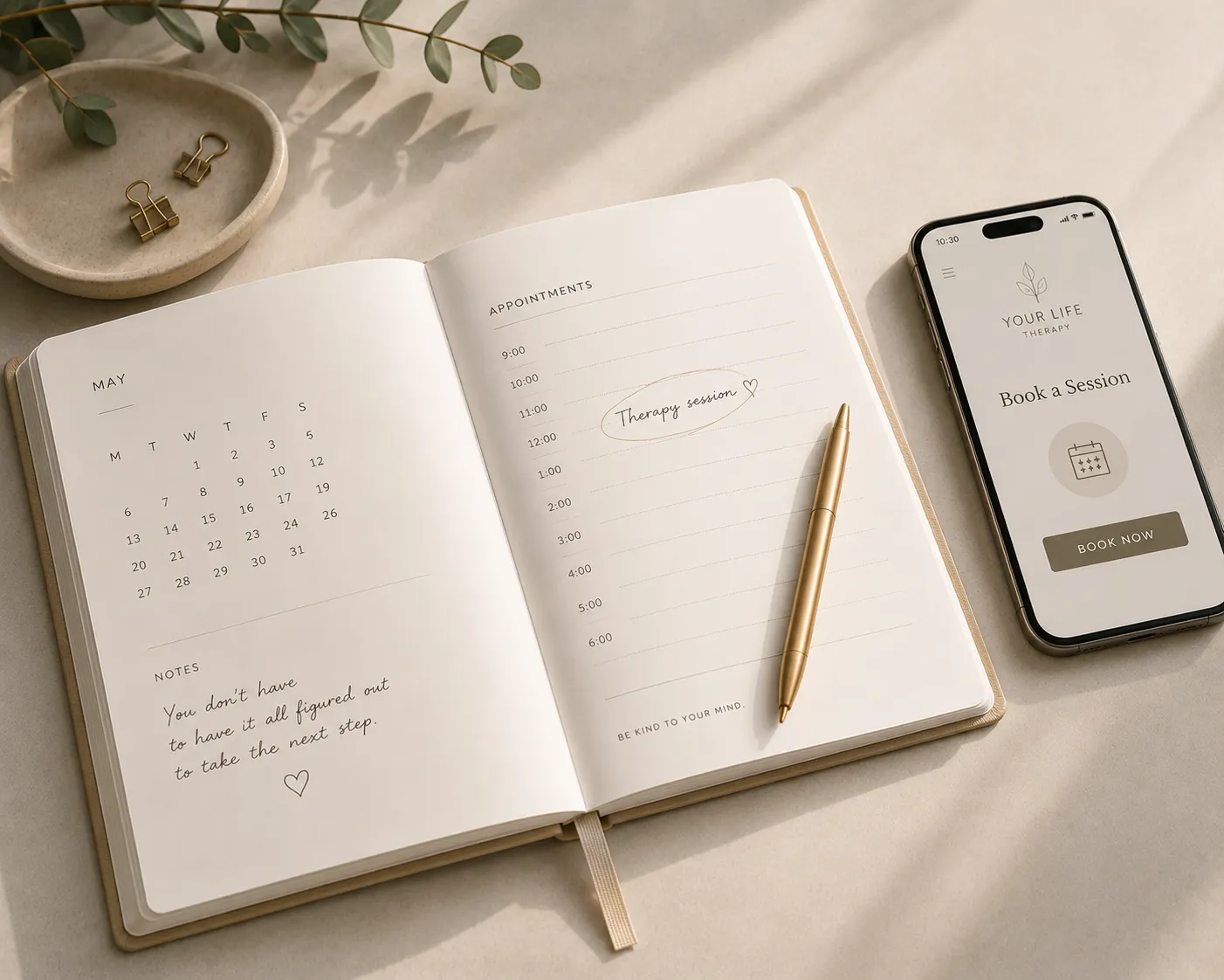 An open planner with a therapy session noted in, a gold pen, and a phone showing the Your Life Therapy booking screen